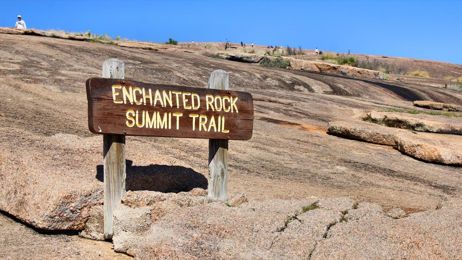 Hikers on the bare pink granite of the Enchanted Rock summit trail under a wide blue sky north of Fredericksburg