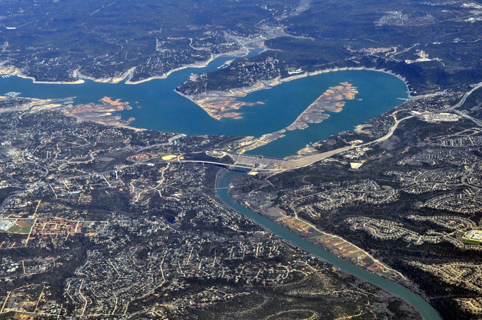 Aerial view of Mansfield Dam and the deep blue main basin of Lake Travis winding into the Hill Country