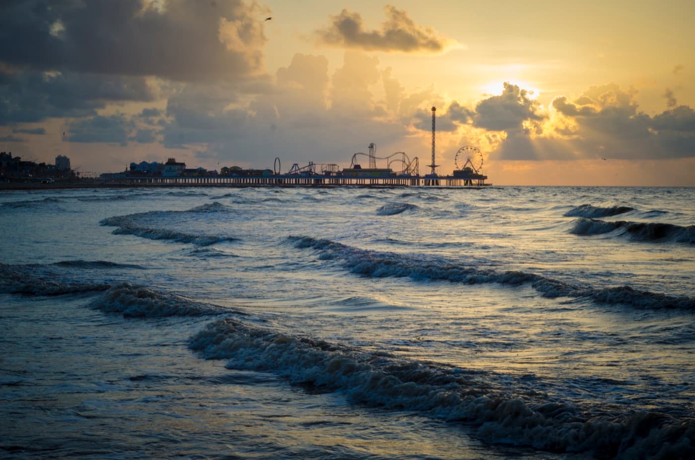 The sun sets over the Gulf surf at Galveston beach, with Pleasure Pier in the distance
