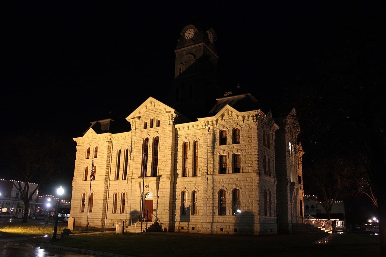 The floodlit limestone Hood County Courthouse and its clock tower at night on Granbury's historic square